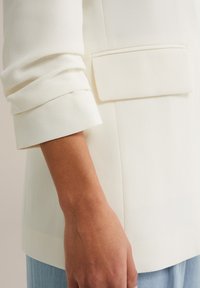 Kremowa marynarka o fakturowanej tkaninie, z podwiniętymi rękawami i naszywaną kieszenią. Krój jest dopasowany, podkreślając jej strukturalny wygląd.