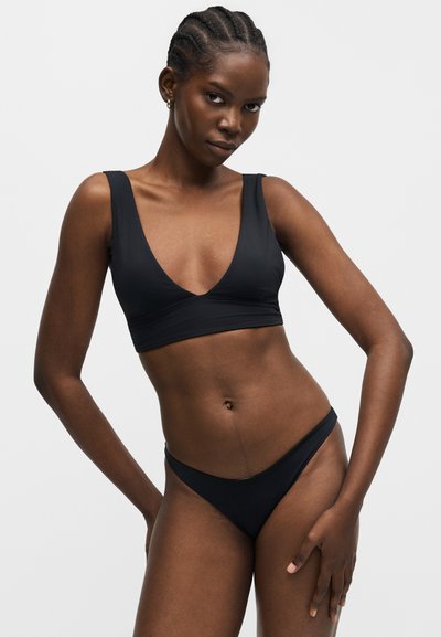 Femme aux cheveux tressés portant un haut de bikini noir avec un profond décolleté en V et un bas assorti sur un fond clair uni.