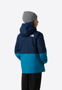 Geacă izolată albastră și verde teal, cu glugă, având un logo pe spate. O căciulă gri tricotată cu nervuri completează ținuta.