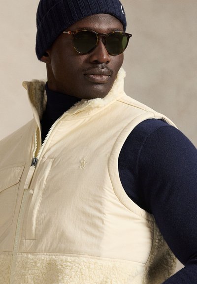 Beige ärmlös väst med en fleeceunderdel, dragkedja framtill och bröstficka; bärs över en marinblå långärmad tröja och kombineras med solglasögon.