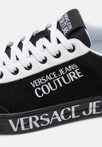 Sorte semskede sneakers med hvite snører, med "VERSACE JEANS COUTURE" branding på siden og en hvit og svart såle.