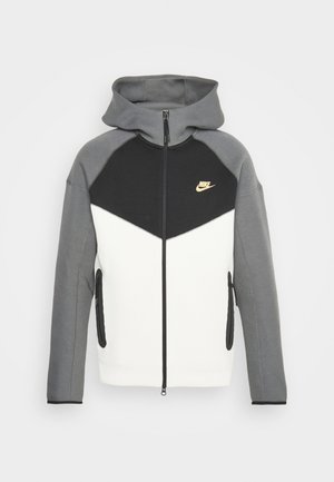 Felpa con zip grigia e nera, con una sezione inferiore bianca, dotata di cappuccio, tasche laterali e un logo Nike dorato sul petto.