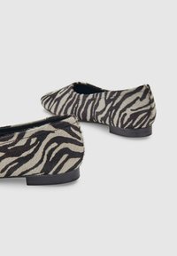 Een paar platte schoenen met een zwart-wit zebraprint patroon en lage zwarte hakken op een effen lichte achtergrond.
