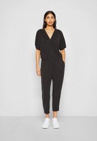 Svart jumpsuit med V-hals, kortermet og elastisk midje. Glatt stoff, smale ben, stylet med hvite sneakers.