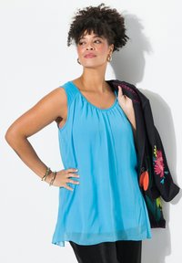 En ärmlös, ljusblå blus med en samlad halsringning och mjuk textur. Modellen håller en svart jacka med färgglad blommig broderi.