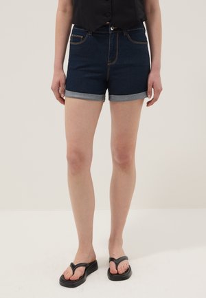 Femme portant un short en jean bleu foncé avec revers roulés, un top noir sans manches et des tongs à plateforme noires, debout sur un sol blanc.