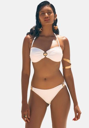 Set bikini bianco con tessuto testurizzato, top bandeau con dettaglio in oro e gambe abbinati con accenti dorati ai lati.