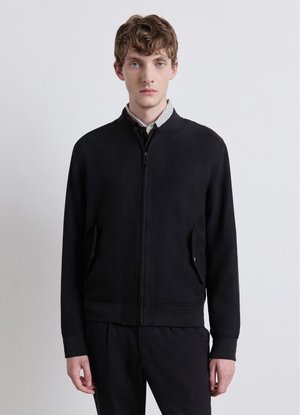 Young man with light brown hair wearing a black zip-up jacket over a collared shirt against a plain light background.
