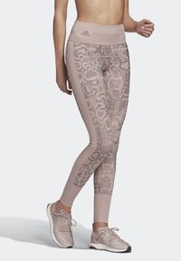 Žena v růžových legínách s vysokým pasem a hadím potiskem a sladěných růžových teniskách, stojící s jednou nohou překříženou vpředu.