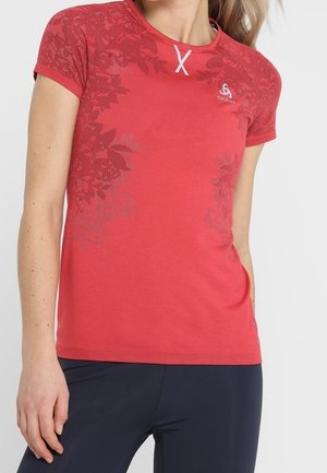 Vrouw draagt een rood T-shirt met korte mouwen met bloemmotieven en een donkere broek, staand met één hand bij haar heup tegen een effen achtergrond.