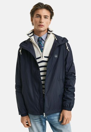 Giacca impermeabile blu navy con cappuccio, dotata di zip, cordini e patch con logo. Indossata sopra un maglione a righe bianche e blu navy.