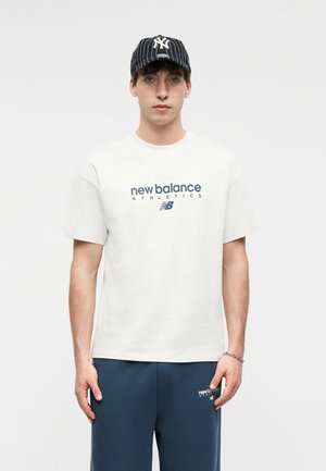 Młody mężczyzna w białej koszulce New Balance Athletics, granatowych dresach i czarnej czapce z paskami New York Yankees, stojący na białym tle.