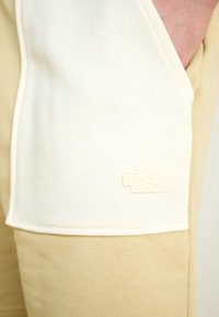 Beige joggebukser med et panel i kremfarge. Funksjoner inkluderer en dekorativ tekstur og en preget logo nederst på panelet.