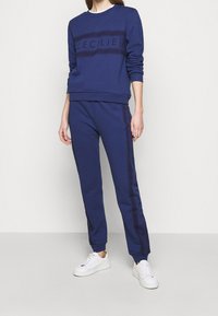 Completo felpa blu navy e pantaloni jogger con accenti testurizzati, con la parola "CECLIE" in evidenza sulla parte anteriore e laterale. Sneakers bianche.