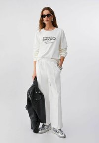 Femme en sweat-shirt blanc et pantalon tenant une veste en cuir noire, portant des baskets argentées, des boucles d'oreilles épaisses, des bracelets et des lunettes de soleil noires.