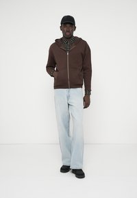 Barna, cipzáras kapucnis pulóver kapucnival, kockás ing fölött viselve. Világoskék farmer és fekete cipő egészíti ki az öltözéket. Egyszerű, hétköznapi kialakítás.