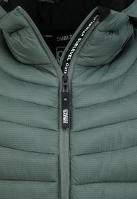 Hellgrüne Pufferjacke mit gesteppter Textur, ausgestattet mit einem Reißverschluss vorne, schwarer Innenauskleidung und Logodetail am Reißverschluss-Zipper.