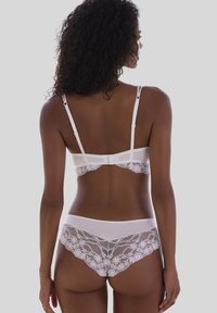 Naine nähtuna seljaga, kandes valget pitsist rinnahoidjat ja sobivaid pitsalaseid aluspükse halli tausta ees.