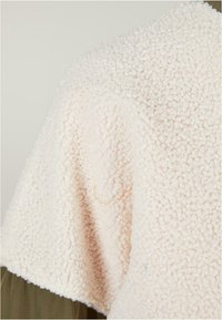 Un tissu en polaire crémeux et duveteux forme le corps principal, avec une surface texturée. Un petit fil orange est visible sur le côté droit.