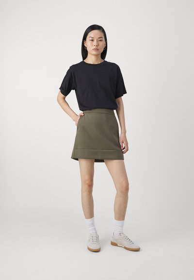 Svart t-skjorte med korte ermer, sammen med et grønt A-linje skjørt. Modellen har på seg hvite sokker og lette sneakers, og står mot en ensfarget bakgrunn.