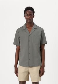 Camisa de manga corta en tejido gris texturizado con collar de muesca, cierre frontal de botones y un bolsillo en el pecho, combinada con pantalones cortos beige.