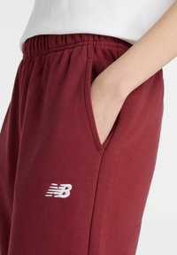 Burgundy sweatpants tillverkade av mjuk bomullsblandning, med elastisk midja, sidofickor och en vit broderad New Balance-logotyp.
