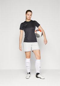 Schwarzes Nike-Shirt, graue Shorts, weiße Socken mit Polsterung und Fußballschuhe. Modell hält einen schwarz-weiß gemusterten Fußball.