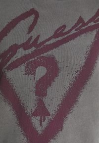 Point d’interrogation violet à l’intérieur d’un triangle avec un texte partiellement en italique au-dessus, imprimé sur un tissu gris texturé.