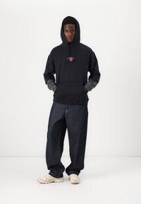 Hoodie bleu marine foncé avec des manches gris, logo rose sur la poitrine. Porté avec un pantalon en denim foncé ample et des baskets de couleur claire. Coupe décontractée.