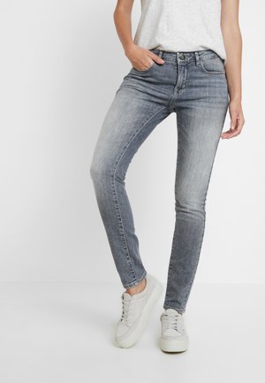 Jean gris délavé slim porté avec des baskets blanches et un haut sans manches gris clair, debout devant un fond blanc uni.