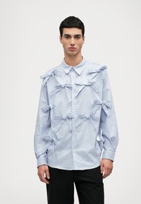 Camisa xadrez azul-claro com vários laços de tecidoelhos na frente. Gola com botões e mangas longas. Corte casual.