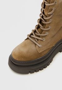 Botín de ante marrón con suela de goma negra gruesa, que presenta ojales metálicos y cordones beige, parte superior texturizada y área de puntera reforzada.