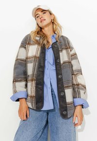 Veste en polar à carreaux gris, blanc et marron, avec un bord en velours côtelé et cinq boutons, associée à une chemise bleu clair et un jean en denim.