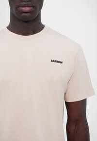 Camiseta de color beige claro hecha de algodón suave, con cuello redondo y mangas cortas, con un pequeño logo negro de "BARROW" en el pecho.