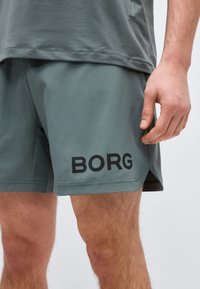 Άνδρας που φοράει γκρι αθλητικό σορτς με την επιγραφή "BORG" στο αριστερό πόδι και ταιριαστό γκρι μπλουζάκι, στέκεται μπροστά σε απλό φόντο.