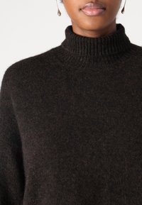 Ruskea neulottu villapaita, jossa korkea resorikaulus. Kangas näyttää pehmeältä ja hieman teksturoidulta.