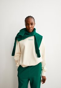 Cremiges Sweatshirt mit grünem Logo, kombiniert mit grünen Jogginghosen. Grüner Schal über die Schultern drapiert. Glatter Stoff und lockere Passform.