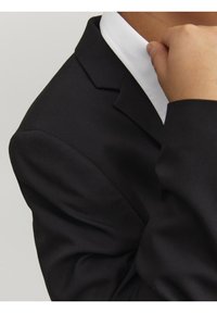 Svart kavaj med strukturerad axel, detaljerad kavajkrage och en vit skjortkrage som tittar fram under. Slät tygtextur.