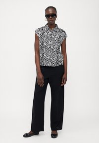 Μπλούζα κοντού μανικιού με γιακά σε μαύρο και λευκό σχέδιο φύλλου, συνδυασμένη με μαύρα παντελόνια φάρδους. Αξεσουάρ περιλαμβάνουν γυαλιά ηλίου και επίπεδα παπούτσια.