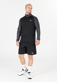 Man som bär en svart och grå zip-up idrottströja, svarta shorts, vita strumpor och vita sneakers står mot en vit bakgrund.