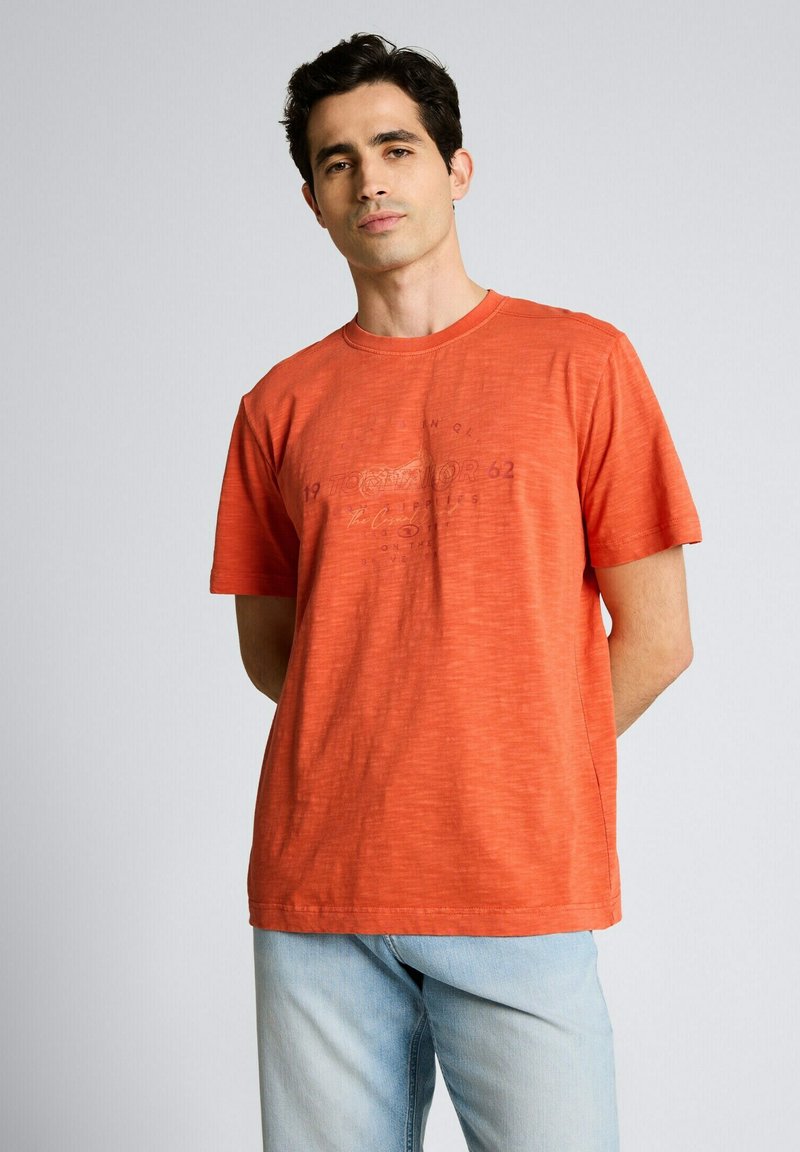 Homme aux cheveux foncés portant un T-shirt orange à manches courtes et un jean bleu clair, debout avec les mains derrière le dos sur un fond uni.