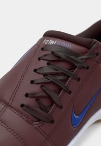 Μπορντό δερμάτινο αθλητικό παπούτσι με σκούρες καφέ κορδόνια. Διαθέτει μπλε λογότυπο Nike swoosh και υφή στην επιφάνεια. Στρογγυλεμένη μύτη και λευκή σόλα.