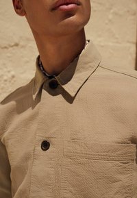 Beige Hemd mit strukturierter Oberfläche, ausgestattet mit einem Kragen, zwei Knöpfen und einer Brusttasche. Der Stoff weist ein dezentes Muster auf.