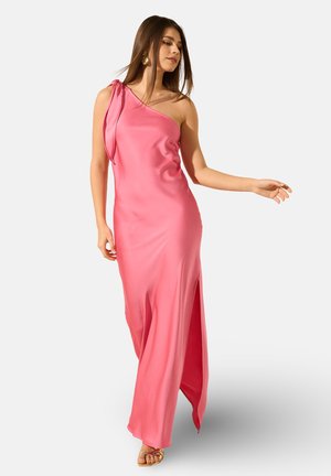 Femme portant une robe en satin rose une épaule avec une fente latérale et des sandales dorées, posant avec le bras droit tendu.