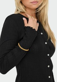 Czarna, prążkowana kardigan z długim rękawem i białymi guzikami, z falbaniastymi mankietami. Na nadgarstku wisi złoty bransoletka.