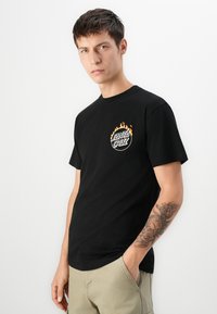 Ung mand med kort, mørkt hår iført en sort Santa Cruz T-shirt med flamme-logo og beige bukser, som viser en tatoveret underarm, hænderne i lommerne.