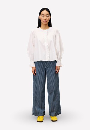 Femme se tenant debout, portant un chemisier blanc à volants, un pantalon large rayé bleu et blanc, et des baskets jaunes vives, sur un fond uni.