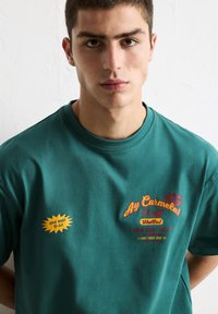 T-shirt en coton vert avec des manches courtes. L'avant présente un texte multicolore et des détails graphiques, y compris une bulle de dialogue avec "Procurez-vous le vôtre !"