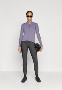 Cykeloutfit bestående af en tætsiddende, lynlås-lavendel top og mørke leggings med en sidelomme. Sort hjelm i hånden.