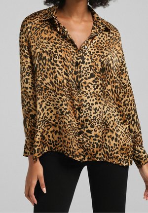 Kvinde iført langærmet, knappet bluse med leopardmønster kombineret med sorte bukser, vist fra brystet til hoften mod grå baggrund.